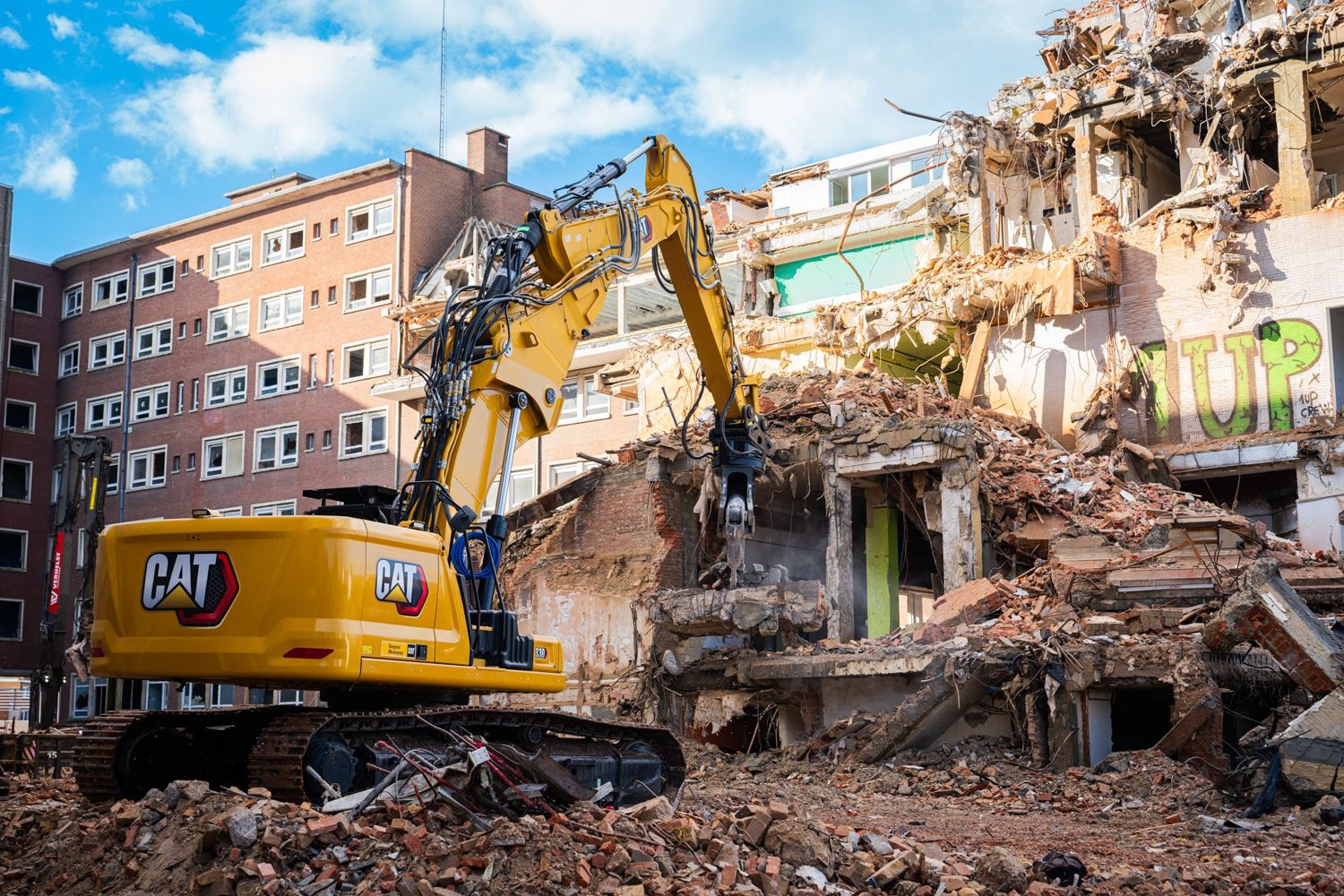 Excavator with hydraulic breaker demolishing a building beside an occupied structure